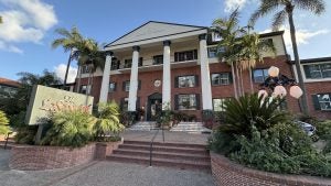 The grand front entrance of The Lafayette Hotel in San Diego featuring a red-brick colonial-style building with white columns, green shutters, and lush palm trees lining the walkway.